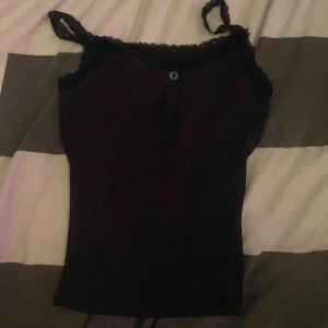 Svart linne med spetsdetaljer - Snygg svart linne med spetsdetaljer vid axelbanden och en liten knapp framtill. Perfekt för en stilren look.