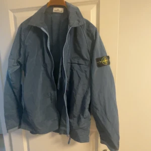 Blå jacka från Stone Island - Säljer en blå jacka från Stone Island med dragkedja och en ikonisk patch på ärmen. Jackan har en avslappnad passform och är perfekt för vår och höst. Den har långa ärmar och en stilren design.