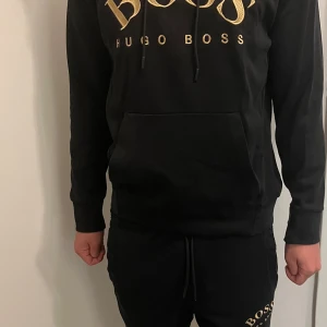 Hugo boss Set, hoodie & byxor - Snygg svart hoodie från Hugo Boss med guldtryck på bröstet. Tröjan har en stor ficka framtill och långa ärmar. Pris kan diskuteras vid snabb affär. Storlek S men passar M oxå, koftan kostar 700kr separat, byxorna kostar 750kr separat, hela setet för 1200kr 
