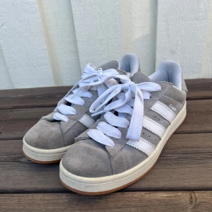 Grå Adidas Campus sneakers - Säljer ett par stilrena grå Adidas Campus sneakers med vita ränder och snörning. Skorna har en klassisk design med rund tå och en bekväm sula i gummi. Perfekta för en avslappnad stil. Storlek 42 & hyfsat bra skick, nypris 1300. 