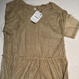 Guldglittrig klänning - Säljer en glittrig guldklänning med korta ärmar och rund hals. Perfekt för festliga tillfällen med sin skimrande yta. Klänningen har en bekväm passform och ett elegant fall.