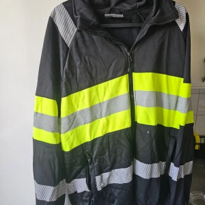Svart och gul jacka från Gesto Workwear - Säljer en svart jacka från Gesto Workwear med gula och grå reflexdetaljer. Jackan har dragkedja framtill och är långärmad. Perfekt för arbete där synlighet är viktigt.