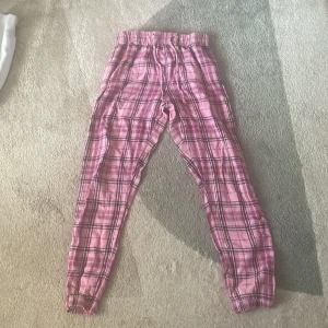 Rosa rutiga byxor med snörning - Säljer ett par rosa byxor med svart rutmönster. Byxorna har en bekväm passform med snörning i midjan och elastiska muddar vid bensluten. Perfekta för en avslappnad stil.