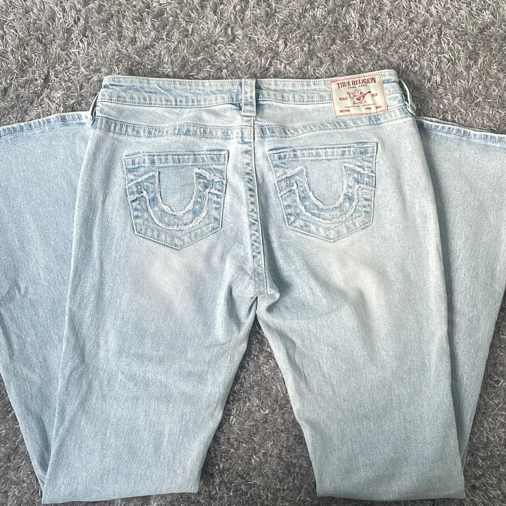 Säljer ett par ljusblå jeans från True Religion med ikoniska bakfickor och broderad logga. De har en låg midja och en bootcut-stil, perfekt för en avslappnad look. Dragkedja och knapp framtill.. Farkut & Housut.
