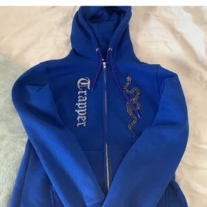 Blå hoodie med dragkedja och ormmönster - Snygg blå hoodie med dragkedja och en cool ormmönsterdetalj på framsidan. Textdetalj i vitt längs ena sidan. Perfekt för en avslappnad stil. Tröjan är ifrån Made By Trapper