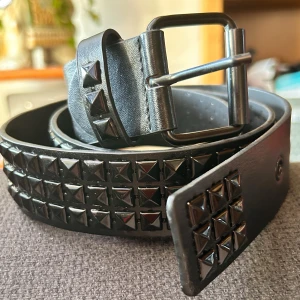 Svart nitbälte vintage/Y2K! - Svart 3-radigt nibälte i vintage/Y2K-stil med svarta pyramidnitar. Längd: 110 cm (85-100 cm innersta till yttersta hålet). Bredd: 4 cm. Oanvänt.