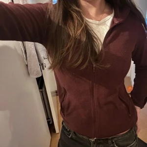 Vinröd fleecejacka med dragkedja - Säljer en mysig vinröd fleecejacka med dragkedja framtill. Jackan har långa ärmar och en bekväm passform, perfekt för kyligare dagar. Den har praktiska fickor på sidorna.