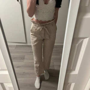 Beige randiga byxor med knytband - Snygga beige byxor med vita ränder och ett knytband i midjan. Byxorna har en avslappnad passform och är perfekta för en stilren look. De är högmidjade och har en croppad längd som ger en modern touch.