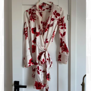 Vit och röd blommig omlottklänning i satin  - Säljer en elegant vit omlottklänning med röda blommor från BikBok i storlek 36. Klänningen har långa ärmar och ett knytband i midjan. 