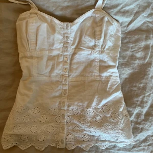 Vit topp med spetsdetaljer - Säljer en söt vit blus med knappar framtill och vackra spetsdetaljer längst ner. Blusen har smala axelband och perfekt till sommaren! Priset går att diskuteras:)