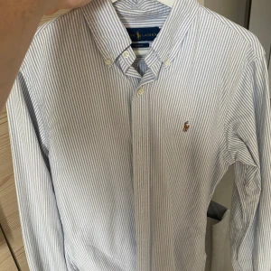 Randig skjorta från Ralph Lauren - Säljer en stilren blå- och vitrandig skjorta från Ralph Lauren. Skjortan har en klassisk krage med knappar och det ikoniska broderade logotypen på bröstet. Perfekt för en elegant look.