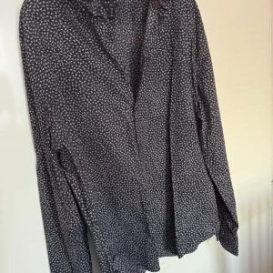 H&M - Snygg mörkblå skjorta från H&M med ett diskret vitt och rött mönster. Skjortan har långa ärmar och knappar framtill. Perfekt för en stilren look.