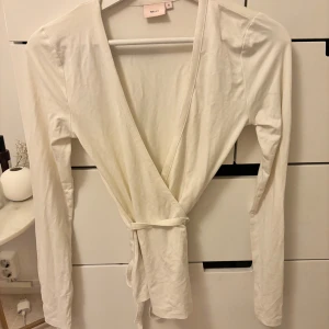 Vit omlottopp från Nelly - Säljer en stilren vit omlottopp från Nelly. Toppen har långa ärmar och ett knytband i midjan för en smickrande passform. Perfekt för en elegant och avslappnad look.