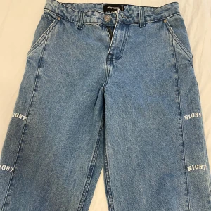 Blå jeans med textdetaljer - Snygga blå jeans med textdetaljer längs benen. De har en klassisk femficksdesign och knappgylf. Perfekta för en avslappnad stil.