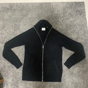 Svart ribbad kofta från Jack & Jones - Säljer en stilren svart ribbad kofta från Jack & Jones med dragkedja framtill. Perfekt för kyliga dagar med sin långa ärmar och bekväma passform. Ett måste i garderoben för en klassisk look. Stor i storlek så passar S.