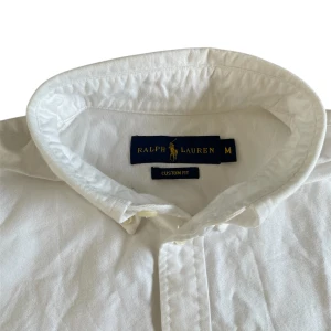 Vit skjorta från Ralph Lauren - Säljer en klassisk vit skjorta från Ralph Lauren i storlek M. Skjortan har en broderad logga på bröstet och knappar framtill. Perfekt för en stilren look.