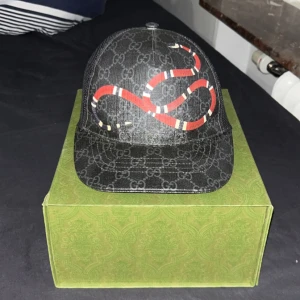 Svart Gucci keps med ormmönster - Snygg svart keps från Gucci med ett iögonfallande rött ormmönster på framsidan. Kepsen har ett diskret GG-mönster och justerbar rem baktill med röd och blå rand. Perfekt för att ge din outfit en lyxig touch. använd men bra skick Pris kan diskuteras. står ej för frakt, kan mötas upp i GBG