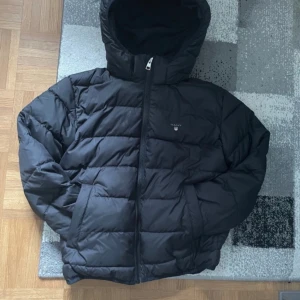 Svart pufferjacka från GANT - Svart GANT-jacka för barn (146/152 cm) Stilren och praktisk svart jacka från GANT i barnstorlek 146/152 cm. Jackan har en klassisk design med diskret logga på bröstet, hög krage och dragkedja framtill. Tillverkad i slitstarkt material som passar både vardag och utomhuslek. Perfekt för mellansäsong med lätt vaddering och bekväm passform.