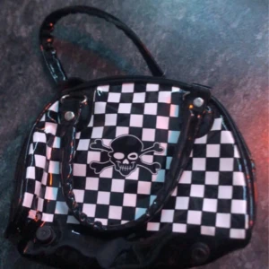 Svart och vit rutig handväska med dödskallemotiv - Cool handväska med svartvitt rutmönster och ett dödskallemotiv på framsidan. Väskan har en glansig finish och dragkedja upptill. Perfekt för att ge din outfit en edgy touch.