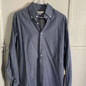 Blå skjorta från J.Lindeberg - Snygg blå skjorta från J.Lindeberg i slim fit. Skjortan har en klassisk krage med knappar och är perfekt för en stilren look. Materialet är en blandning av bomull och syntet vilket ger en skön känsla. skulle säga skjortan är lite mer mot M men passar även folk med S