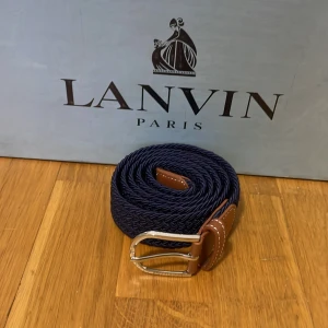 Blått flätat bälte  - Snyggt blått flätat bälte med bruna skinn-detaljer och en stilren silverfärgad spänne. Perfekt för att ge en touch av elegans till din outfit. Passar bra till både jeans och kostymbyxor.