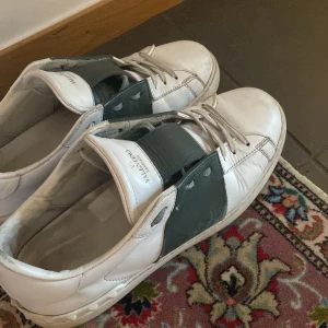 Valentino sneakers i vitt och grönt - Snygga Valentino sneakers i vitt med gröna detaljer. Skorna har snörning och en bred grön rem över vristen för en unik look. Perfekta för en stilren och modern stil.