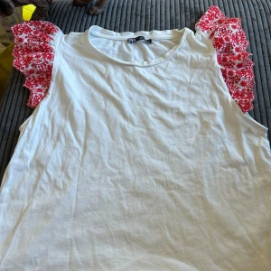 Vit topp med röda blommiga volangärmar från Zara - Säljer en vit topp från Zara med unika röda blommiga volangärmar. Toppen är ärmlös och har en rund halsringning. Perfekt för en somrig look! 🌸 den har en jätte liten fläck och ett jätte jätte litet hål