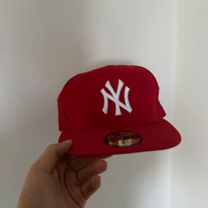Röd keps från New Era - Snygg röd keps från New Era med vit broderad logga framtill. Kepsen har en klassisk design med platt skärm och justerbar passform. Perfekt för att ge din outfit en sportig touch. Aldrig använd