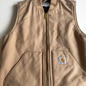 Beige väst från Carhartt - Säljer en snygg beige väst från Carhartt med dragkedja och praktiska fickor. Perfekt för lager-på-lager under höst och vår. Klassisk design med Carhartt-logga på framsidan. Materialet är kanvas, en lite fläck typ längst ner på dragkedjan, annars bra skick
