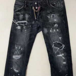 Svarta jeans med slitningar från Dsquared2 - Snygga svarta jeans från Dsquared2 med coola slitningar och en unik dragkedjedetalj vid fickan. Jeansen har en modern look med en tvättad finish och är perfekta för en trendig stil. Jeansen är i jättebra skick, det finns även påse till den och dsq prislappen. Priset kan diskuteras såklart.