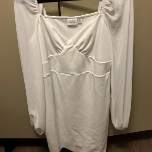 Vit student klänning från bubleroom, märket Vila - Elegant vit klänning från Vila med långa ärmar och en stilren design. Klänningen har en vacker drapering framtill och en smickrande passform. Perfekt för speciella tillfällen.