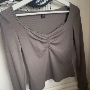 Grå ribbad topp - Säljer en stilren grå ribbad topp med v-ringning och rynkdetalj framtill. Toppen har långa ärmar och en figurnära passform, perfekt för en chic vardagslook.