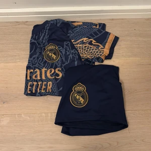 Adidas fotbollströja REAL MADRID - Säljer en mörkblå fotbollströja från Adidas med ett coolt drakmönster i guld samt ett par mörkblåa shorts som ingår. Vini Jr nr 7 på ryggen. Original pris för hela bundle är 899kr från unisport (de säljer inte det längre) passar från 146-150