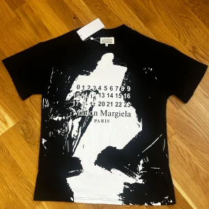 Svart t-shirt från Maison Margiela - Säljer en svart t-shirt från Maison Margiela med ett unikt tryck i vitt på framsidan. Tröjan är i storlek L och är i nyskick med lappen kvar. Hör av er vid frågor och funderingar!