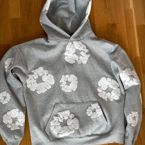 Grå hoodie från Denim Tears - Säljer en grå hoodie från Denim Tears med unika vita blommönster över hela tröjan. Den har en klassisk känguruficka och en bekväm huva. Perfekt för en avslappnad stil. Tillverkad i USA.