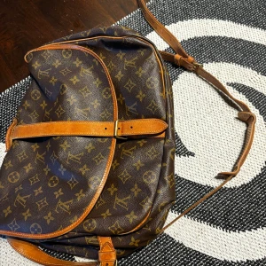 Brun axelväska från Louis Vuitton - Säljer en klassisk brun axelväska från Louis Vuitton med det ikoniska monogrammönstret. Väskan har justerbar axelrem och spänne framtill. Perfekt för att bära dina dagliga nödvändigheter med stil. Jag valde no size men väskan är köpt second hand därav inget kvitto och har den får plats med mycket. Älskar den men använder så mycket. Så jag hoppas den hittar någon som har bättre användning för den. 