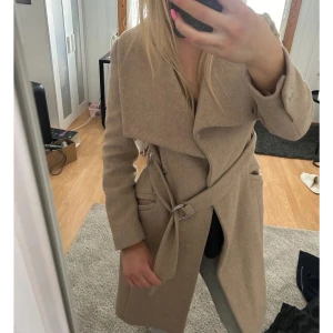 Beige kappa med bälte - Elegant beige kappa med bälte och knappar. Perfekt för höst och vår. Långärmad design med stilren look.