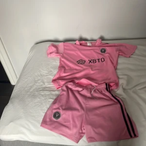 Rosa fotbollströja och shorts från Inter Miami - Säljer en rosa fotbollströja och matchande shorts från Inter Miami. Tröjan är kortärmad med klubbens logga och sponsortryck på framsidan. Shortsen har en elastisk midja och klubbens emblem. Perfekt för fotbollsfans som vill visa sitt stöd för laget.