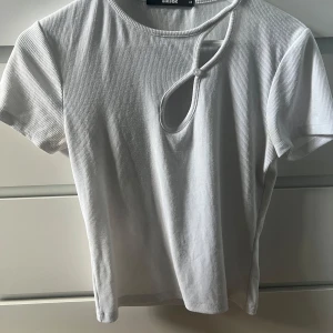 Vit ribbad topp från BikBok - Säljer en stilren vit ribbad topp från BikBok med en unik knapplösning vid halsen. Toppen har korta ärmar och en modern design som passar perfekt till både jeans och kjol. Perfekt för en avslappnad men trendig look!