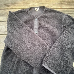 Svart teddyjacka med knappar - Mysig svart teddyjacka med knäppning fram. Perfekt för kyliga dagar med sin mjuka och varma textur. Jackan har långa ärmar och en enkel design som gör den lätt att matcha med olika outfits.