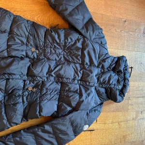 Svart dunjacka från Moncler - Snygg svart dunjacka från Moncler med dragkedja och knappar. Jackan har en quiltad design och en hög krage för extra värme. Perfekt för kyliga dagar. Hör av er för frågor eller funderingar 