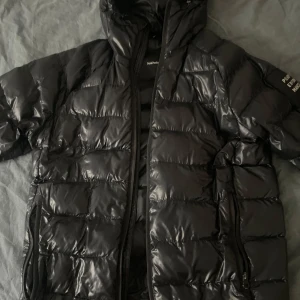 Svart pufferjacka från Peak Performance - Säljer en svart pufferjacka från Peak Performance. Jackan har en glansig yta och är utrustad med dragkedja. Perfekt för kyligare dagar med sin vadderade design.