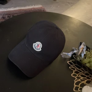 Svart keps från Moncler - Snygg svart keps från Moncler med justerbart band i rött, vitt och blått. Kepsen har en broderad logotyp framtill och är perfekt för en stilren look.100% original!