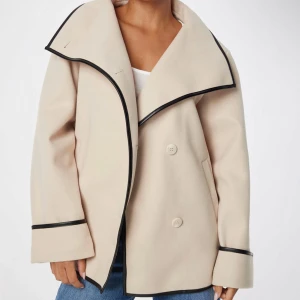 Beige kappa med svarta detaljer - Snygg beige kappa med svarta kantdetaljer och stora knappar. Den har en elegant krage och långa ärmar. Perfekt för en stilren look. Nypris 899kr, säljer för 300kr