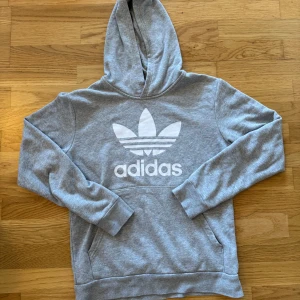 Grå hoodie från Adidas - Säljer en stilren grå hoodie från Adidas med det klassiska logotyptrycket i vitt på bröstet. Tröjan har en bekväm huva och en praktisk magficka. Perfekt för en avslappnad stil. Säljer för att tröjan har blivit för liten för mig 