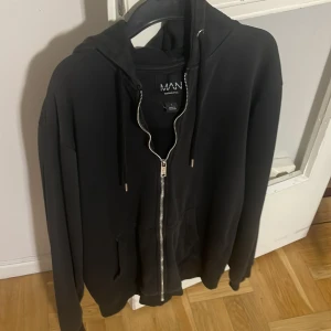 Svart hoodie från boohooMAN - Säljer en stilren svart hoodie från boohooMAN. Den har en dragkedja framtill och en praktisk huva med snörning. Perfekt för en avslappnad look.