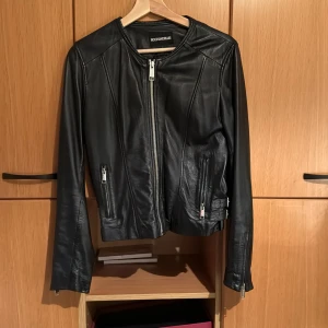 Svart skinnjacka från Rockandblue - Snygg svart skinnjacka från Rockandblue med dragkedja framtill och på ärmarna. Jackan har en stilren design med justerbara spännen i midjan för en perfekt passform. Perfekt för en cool och edgy look.