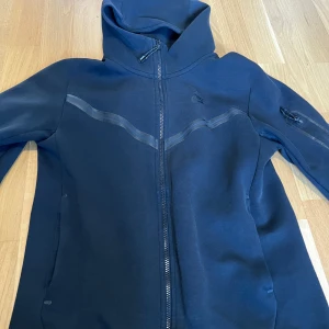 Nike Tech Fleece - Säljer en svart huvtröja från Nike med dragkedja framtill. Tröjan har en stilren design med långa ärmar och en praktisk huva. Perfekt för en sportig look eller avslappnade dagar. Har använt den flera gånger så det finns några noppor! storlek S men passar M också