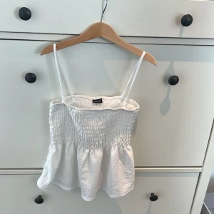 Vit linnetopp från Gina Tricot - Säljer en söt vit linnetopp från Gina Tricot med smala axelband och smockdetaljer. Perfekt för varma sommardagar. Toppen har en luftig passform och är tillverkad i ett mjukt material.
