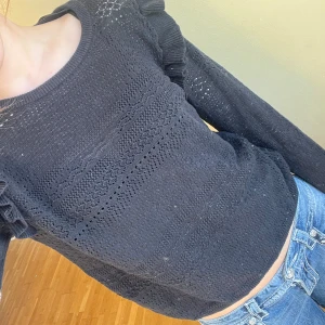 Svart stickad tröja med volangdetaljer - Säljer en svart stickad tröja med fina volangdetaljer på axlarna. Tröjan har ett vackert mönster och långa ärmar. Passar både till jeans och kjol för en stilren look.